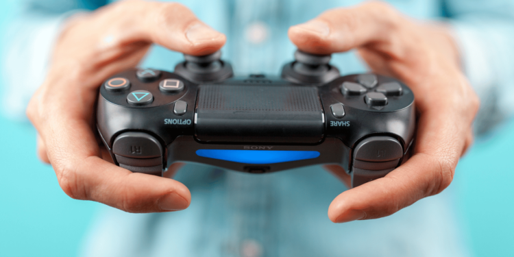 Mãos segurando um controle de PS4, com um fundo azul claro, pronto para jogar no PC.