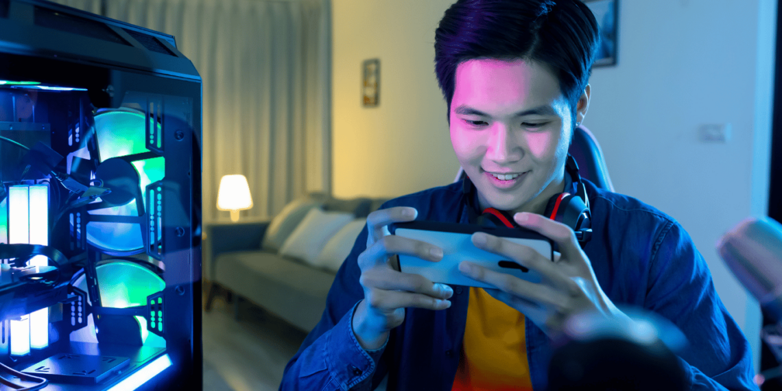 10 Melhores Celulares Gamer Baratos para Você! 1 Pessoa jogando em celular gamer barato ao lado de um setup de PC iluminado, demonstrando a experiência de jogo móvel de alta qualidade.
