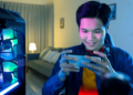10 Melhores Celulares Gamer Baratos para Você! 10 Pessoa jogando em celular gamer barato ao lado de um setup de PC iluminado, demonstrando a experiência de jogo móvel de alta qualidade.